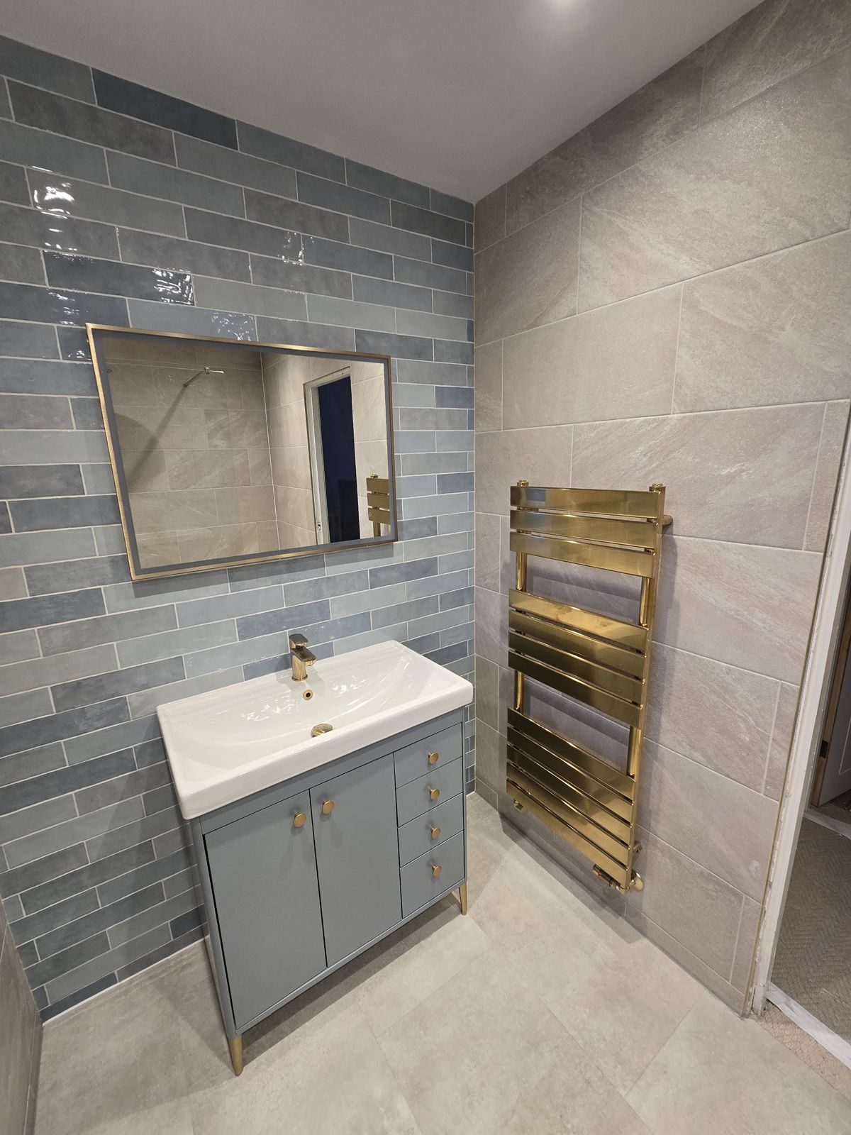 A grey vanity unit and mirror against blue-grey tiles, with a brass towel rail