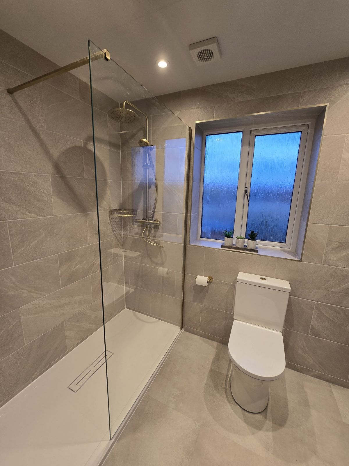 A walk-in shower with a frameless glass panel, brass fittings and large grey tiles